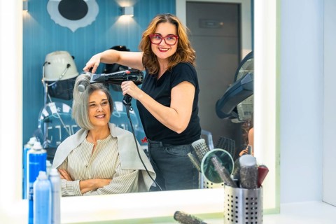 Female hairdresser smiling at camera while blow drying a mature customer's hair in a modern salon,