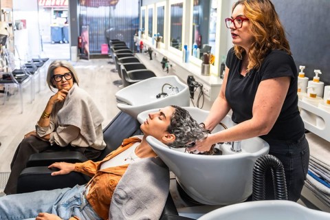 Hairdresser shampooing a young woman at a modern salon while an older client watches nearby,