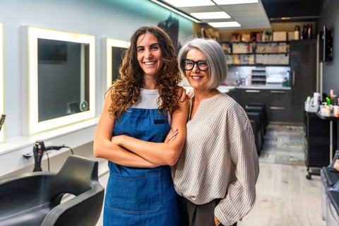 Two cheerful women hairstylists, one younger in an apron and one older with gray hair, standing