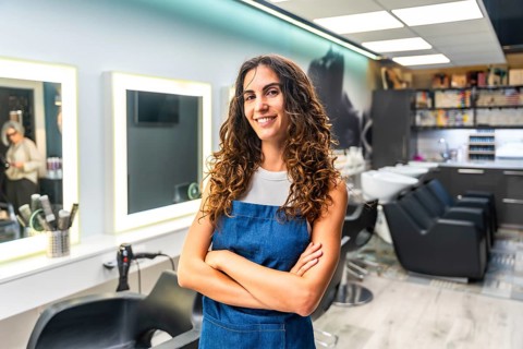Confident young woman with curly hair wearing an apron and smiling, standing with crossed arms in