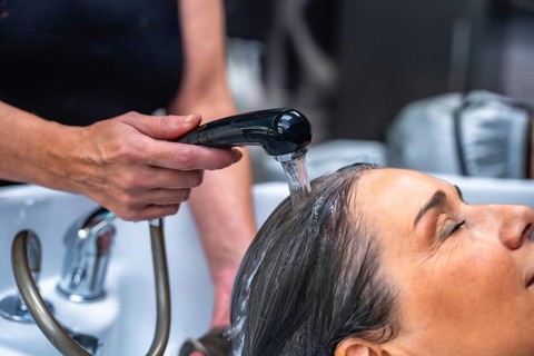 Hairdresser's hands rinsing a woman's hair with water from a shower head at a salon basin, focusing