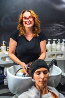 Hairdresser smiling while washing client's long red hair over salon sink, providing professional