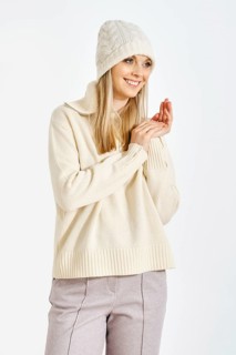 A woman stands with a smile, wearing a sweater and a hat. She clasps her hands together in front of