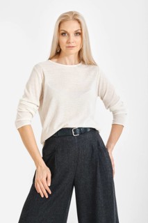 A woman stands with a neutral expression in a studio. She wears a light top and gray wide pants.