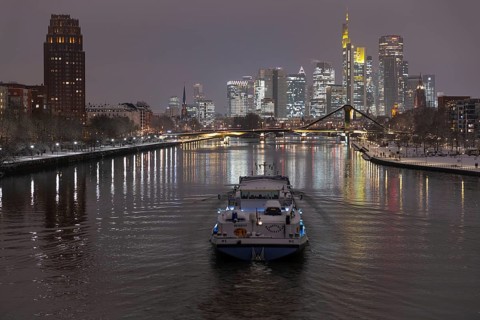 A ship sails towards the snow-covered and glowing Frankfurt banking skyline in the evening,