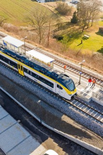 A train is standing on the platform, people on the track, surrounded by fields and countryside,