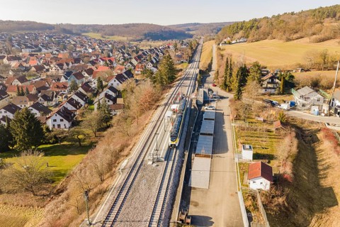 Railway line runs through a village with red-roofed houses, surrounded by trees and meadows, test
