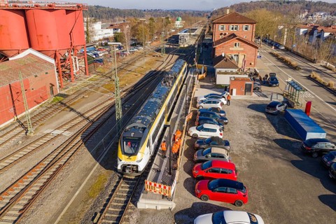 Train stands at a train station next to parked cars and industrial buildings, several workers in