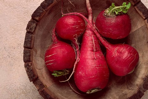 A collection of vibrant red radishes sits in a round wooden bowl, showcasing their natural texture
