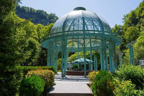 An elegant glass and metal pavilion in a green, sunny landscape surrounded by trees, pavilion with