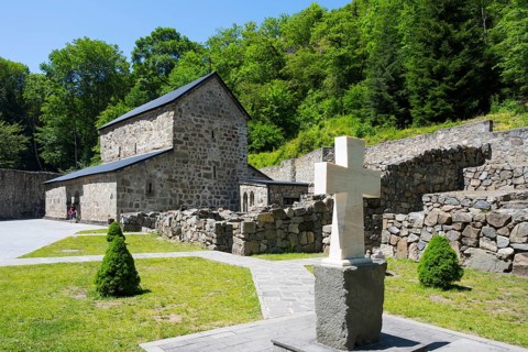 Stone cross in front of a historic church surrounded by green nature, Chitakhevi Church of St.