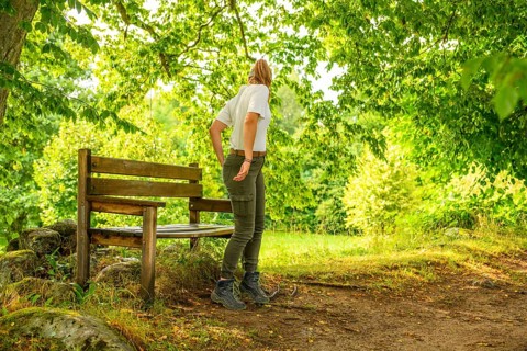 Young woman in summer clothes standing next to a wooden bench in sunny forest, Linnés Råshult,