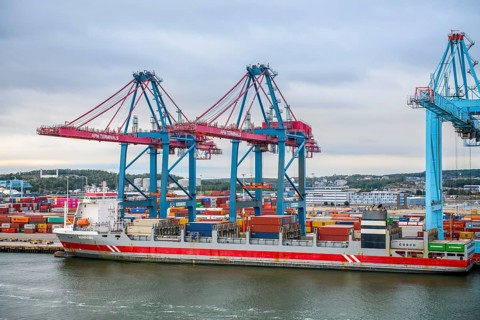 Large port with several blue and red cranes and container ships under cloudy sky, Gothenburg,