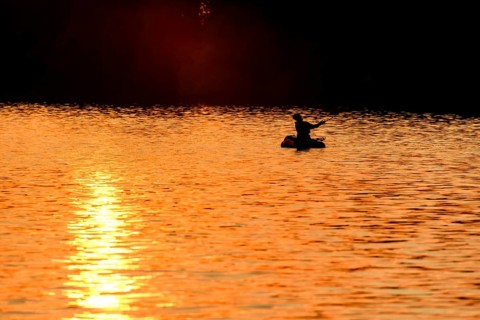 Angler sport fisherman in his belly boat in orange-red lake at sunset, silhouette in calm water,