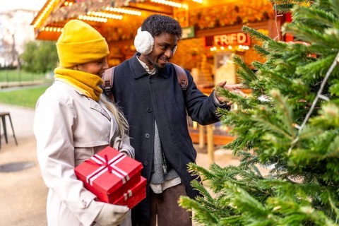 Diverse couple holding christmas gifts and touching a decorated pine tree at a vibrant winter