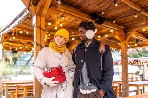 Young diverse couple walking together at a christmas market or winter fair, holding red gift boxes