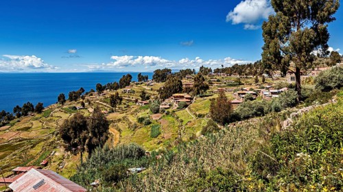 Village with huts on a hill surrounded by green fields and a blue lake under clear skies, the