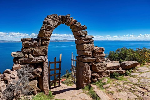 Picturesque stone arch with wooden gate opens to a calm, deep blue lake under clear skies, stone
