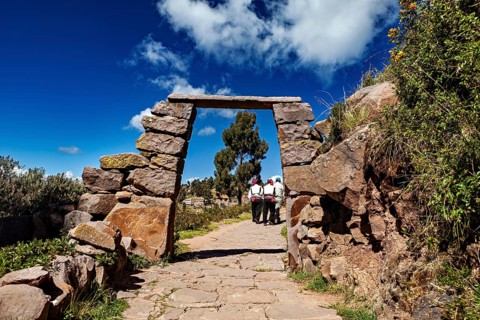 People wearing traditional clothes walk under a rectangular stone arch on a rural path, stone gates