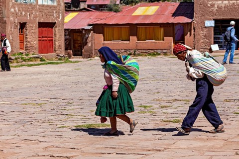 Two woman in colorful clothes carry loads on their backs and walk through a sunny village in