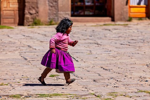 Little girl in purple skirt and pink sweater walks across a paved square, children from Taquile