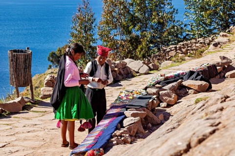 People wearing traditional clothing sell goods on a rock path, overlooking a large lake, the woman