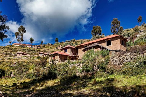 Houses cluster on a hillside under a dramatically cloudy sky framed by trees and vegetation, The