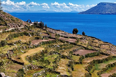 Terraced fields stretch down to a clear blue lake, framed by stone arches and hills, the