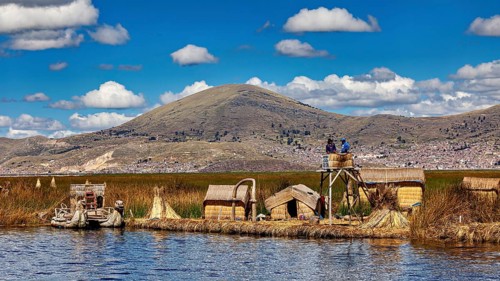 Reed huts and lakeside observation tower with mountains in the background under blue sky, The