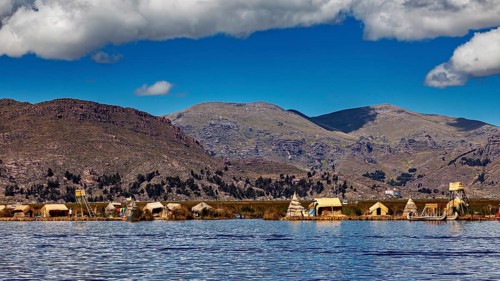 Reed huts on the shores of a tranquil lake in front of a vast mountain landscape under a blue sky,