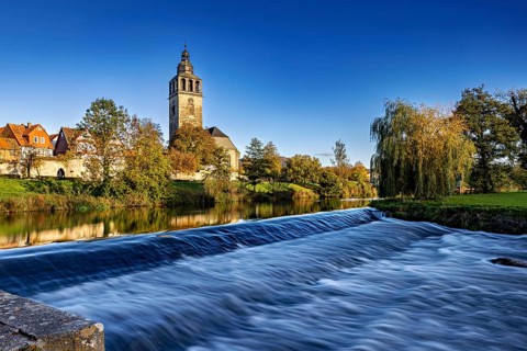A church on the river with a small waterfall in the foreground and autumn trees in the background,