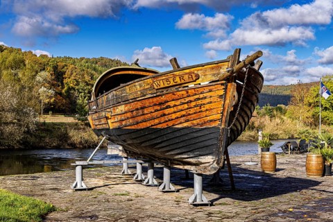 Historic wooden boat on riverbank, surrounded by autumn trees under a clear sky, the port of