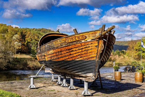 Historic wooden boat on the riverbank on supports, surrounded by autumn trees under a blue sky, the