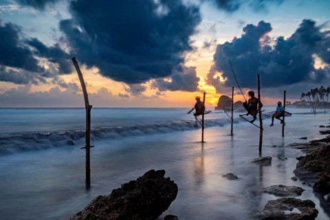 Silhouette of fishermen at sunset over the blue and orange sea, Koggala beach with stilt fishermen
