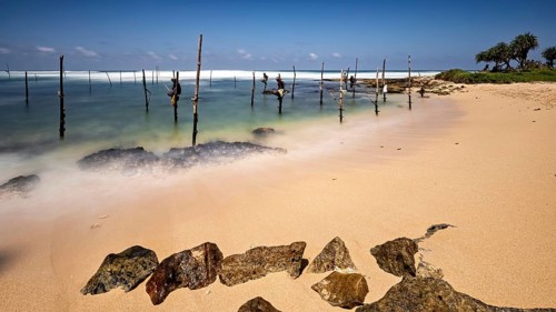 Clear beach with visible rocks and wooden posts in the water, Koggala beach with stilt fishermen in