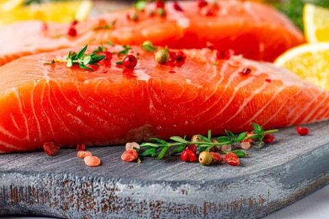 Two pieces of salmon fillet sit on a wooden cutting board. They are topped with herbs and pink