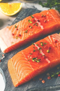 Two pieces of salmon fillet sit on a wooden cutting board. They are topped with herbs and pink