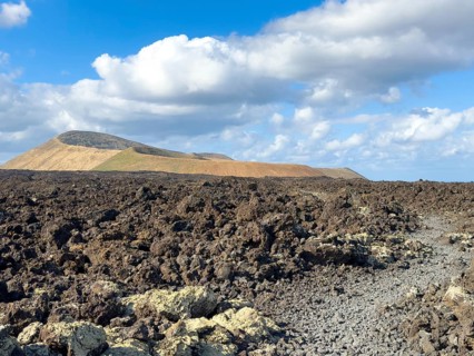 Hiking trail through lava rock to Caldera Blanca volcano against blue sky with white clouds, Mancha