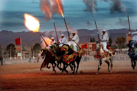 Riders on horses shoot volleys synchronously into the evening sky at Fantasia, El Kelâa m'Gouna,