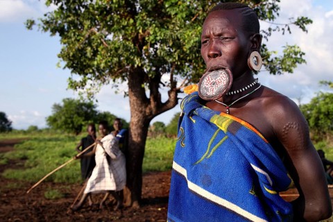 Mursi woman with lip plate in front of a tree in Mago National Park. A village can be seen in the