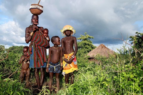 Mursi woman with children in front of a grass hut and cloudy sky, Mago National Park, Ethiopia