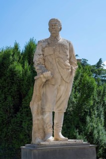 Stone statue of a man in uniform in front of a green hedge outdoors, Stalin monument, sculpture,