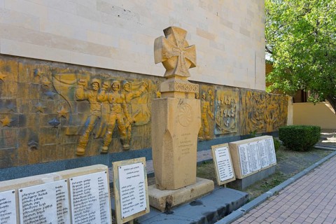 Monument with artistic relief surrounded by memorial plaques in shady surroundings, monument in