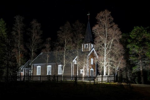 An illuminated church stands at night surrounded by trees and a wooden fence