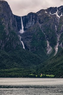 A narrow waterfall plunges from dramatic mountains into a dark fjord
