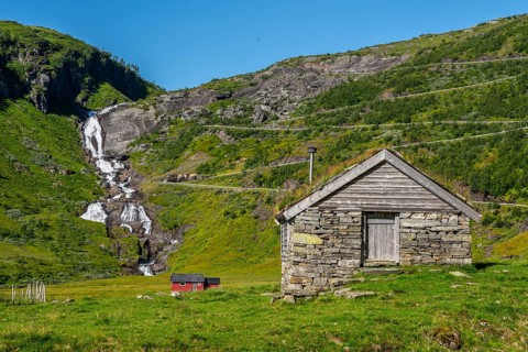 A stone hut stands in front of a rippling waterfall in the middle of green meadows