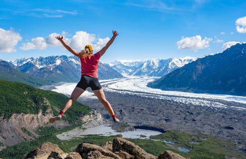 Young woman jumping into the air, view of impressive mountain landscape with Matanuska glacier and