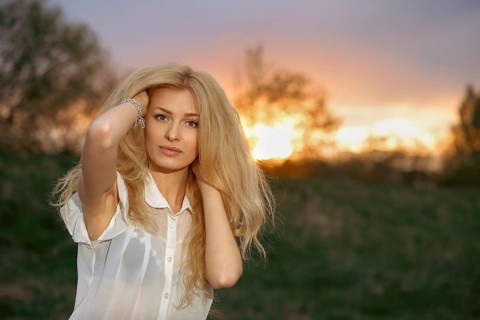 A woman stands in a field with blonde hair flowing as she poses during sunset. The sun creates a