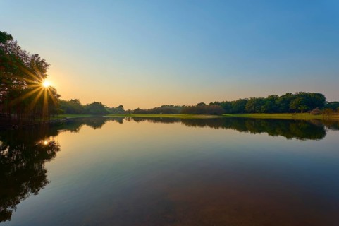 The first rays of sunshine fall on the calm lake, surrounded by green trees and clear skies,