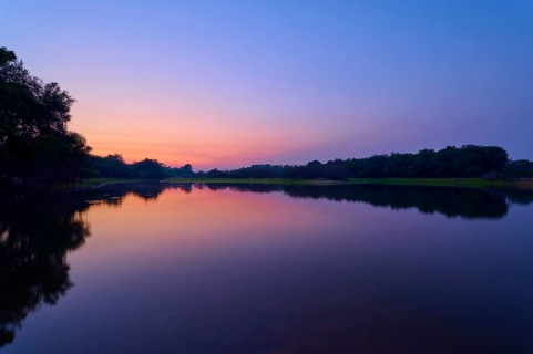 The lake reflects the purple and pink morning sky, framed by dark tree lines, Fazenda Barranco
